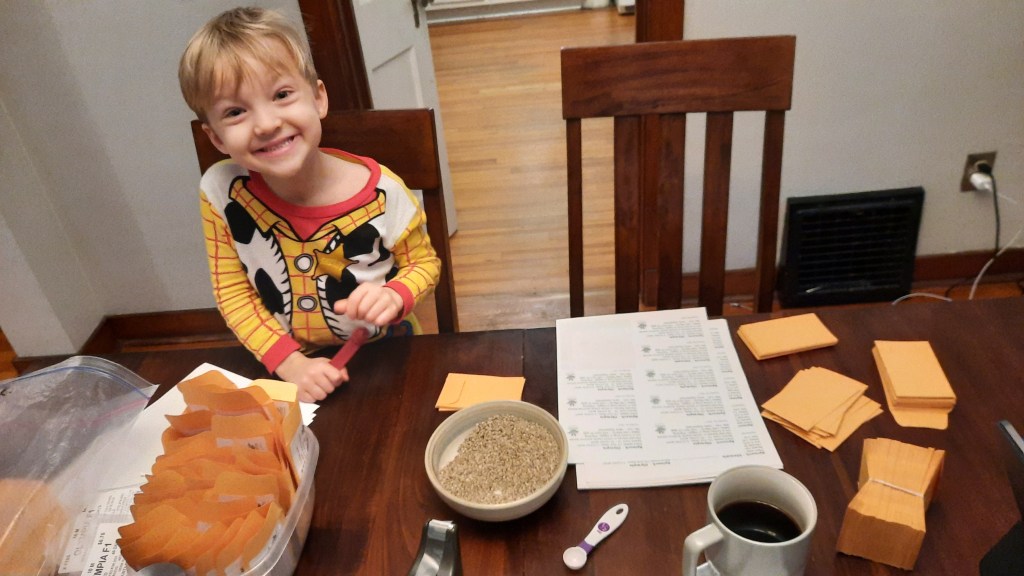 A young boy smiles at a table full of bulk seeds, labels and seed envelopes.