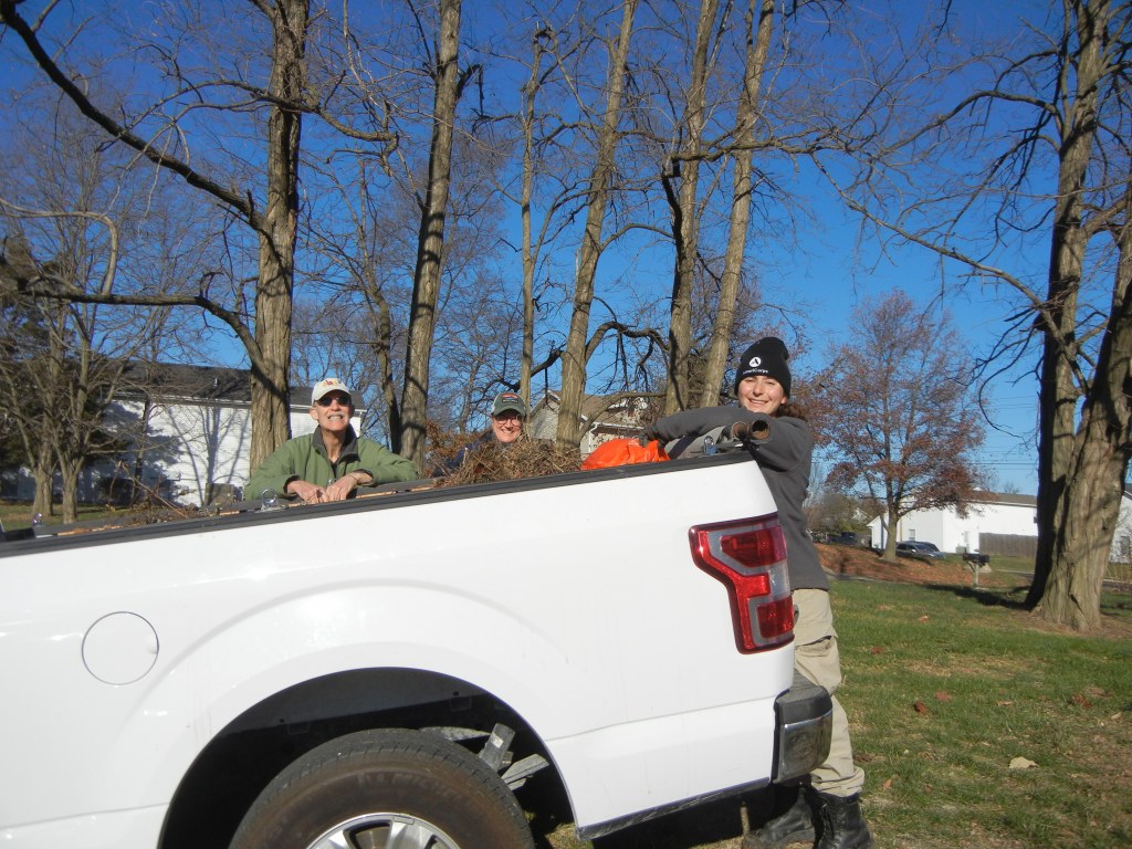 Three volunteers look at the camera as they lean over the back of a pickup full of garden debris.