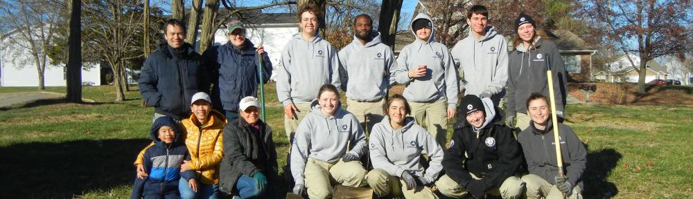 Americorps volunteers and garden leaders pose kneeling and standing in two rows in an open area with grass in front and bare trees behind them.