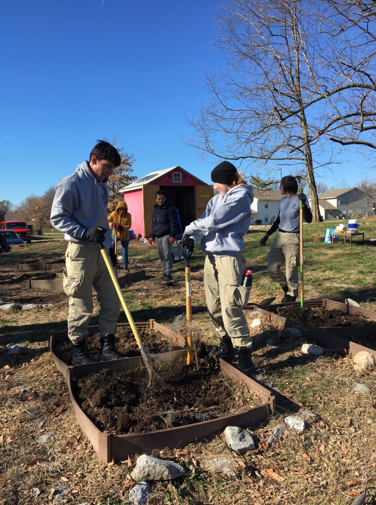 Americorps volunteers spread compost in low raised garden beds at a community garden on an early winter day. A shed with a solar panel is in the background.