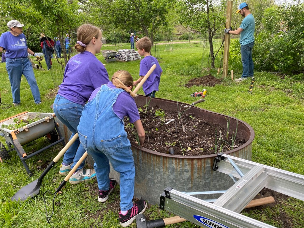 Young For Columbia volunteers clear a small raised metal bed in the foreground while a man works on putting up deer fencing in the background at Interfaith garden.