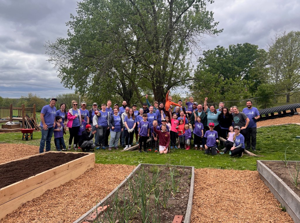 A large group of children, staff and For Columbia volunteers poses in the yard at Windsor Street Montessori with raised garden beds and mulched areas in the foreground.