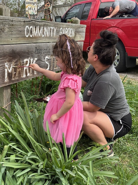 A young child and her mother repaint a sign that says "Community Garden sponsored by Mark Stevenson"