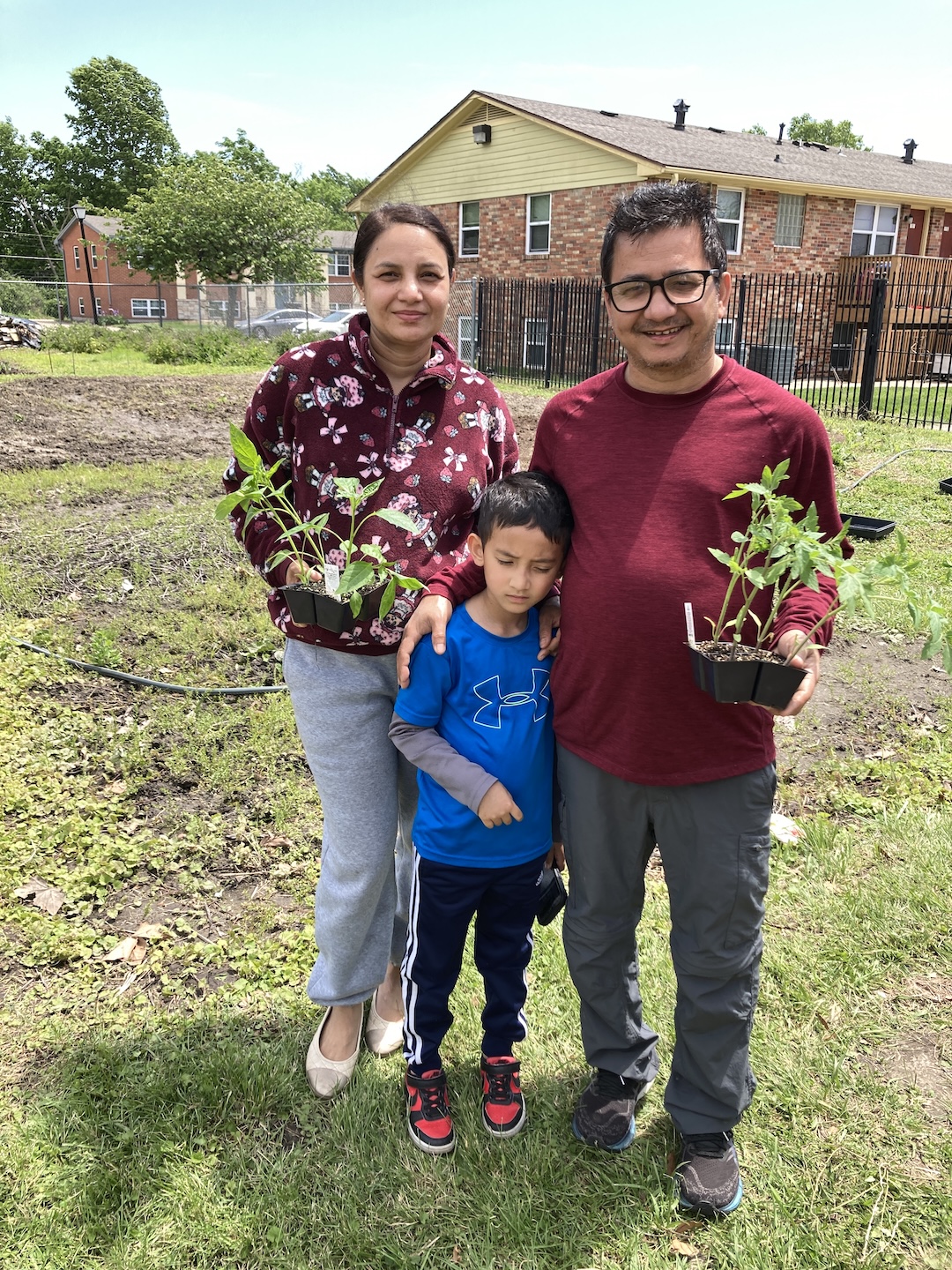 A pair of adults with a child stand in a grassy path besinde community garden plots, holding plant seedlings